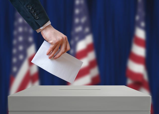 Election in United States of America. Voter holds envelope in hand above vote ballot. USA flags in background. Democracy concept.