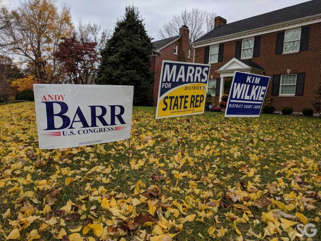 3 political signs in a yard with yellow leaves on the ground and a house in the background