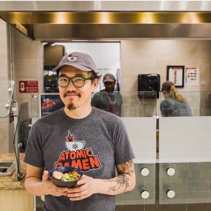 a man in a gray shirt that says atomic ramen smiling at the camera with a hat on