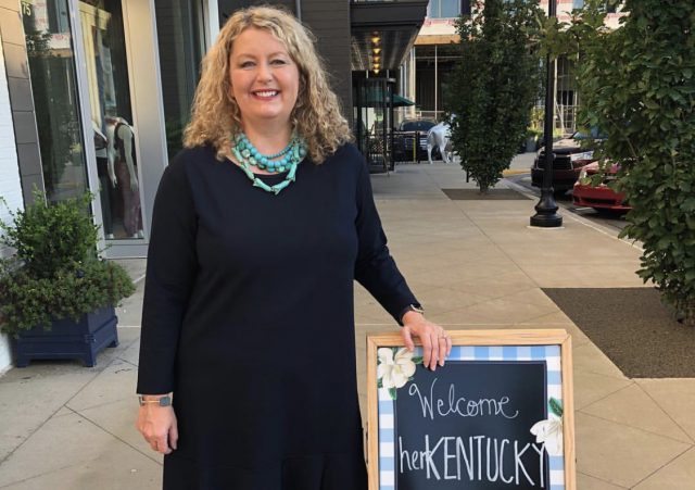 benefiting: a woman in a black dress with a turqoise necklace standing next to a sign that says welcome herkentucky