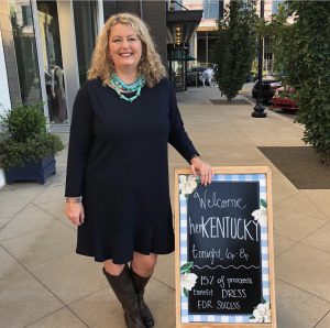 shop & sip: a woman in a black dress with a turqoise necklace standing next to a sign that says welcome herkentucky