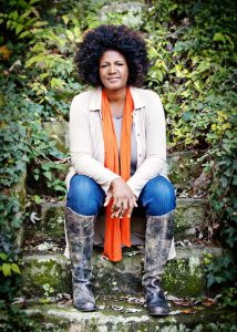 Ouita Michel: woman in a jacket and scarf sitting in nature smiling at the camera